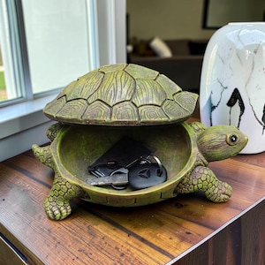 May include: A green turtle-shaped trinket box with an open lid, revealing keys inside. The box is on a wooden surface, with a decorative vase in the background. The turtle has detailed shell and skin textures.