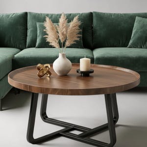 Puede incluir: Una mesa de centro redonda de madera con acabado de veta marrón oscuro y borde elevado, sostenida por un marco de metal negro. Sobre la mesa hay un jarrón de cerámica blanca con hierba de la pampa beige, una escultura geométrica dorada y una vela color crema.