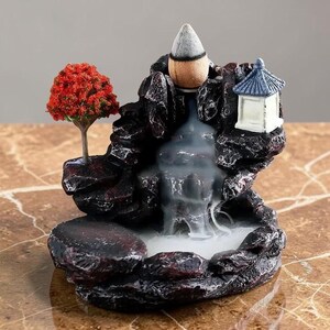 May include: A dark brown rock-shaped incense burner featuring a small red-leafed tree on the left, a white pagoda structure on the right, and a cone-shaped incense piece at the top, with white smoke cascading down like a waterfall into the base.