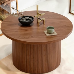 May include: A round coffee table with a wood grain top and a cylindrical base featuring vertical slats. On the table sits a black bowl filled with smooth dark stones and a sprig of greenery, a gold-toned geometric sculpture, and a small green cup.