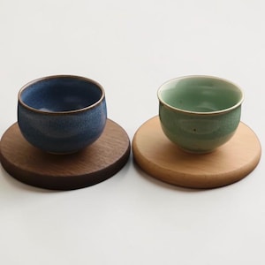 以下が含まれることがあります： 深い青色の斑点模様の釉薬がかかった小さな陶器のティーカップと、淡いミントグリーンのひび割れ模様の釉薬がかかったティーカップが、それぞれ円形の木製コースターの上に置かれています。コースターは濃い木の色と明るい木の色で構成されています。