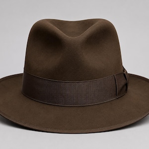 Puede incluir: Un sombrero fedora de color marrón oscuro que presenta una copa estructurada con un pliegue central y un frente pellizcado. El sombrero incluye una cinta de grosgrain marrón oscuro a juego que rodea la base de la copa, con un pequeño lazo lateral.