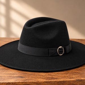 Könnte beinhalten: Ein schwarzer Fedora-Hut mit breiter Krempe und strukturierter Krone. Der Hut verfügt über ein schwarzes Band, das die Basis umschließt und an der Seite mit einem kreisförmigen, goldfarbenen Metallring verziert ist, was ein einheitliches Design ergibt.