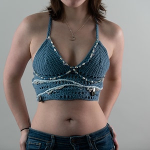 May include: A blue crochet halter top with a V-neck design. The top is adorned with white and silver embellishments along the neckline and lower edge. The top is worn with blue jeans.