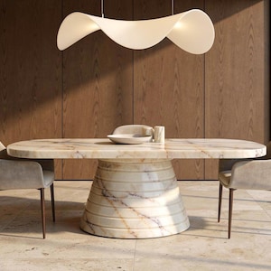 May include: A modern dining room set featuring a marble oval table with a unique, layered base. Two upholstered chairs with wooden legs flank the table. A white, ribbon-like light fixture hangs above. The room has wood paneling and a neutral color palette.