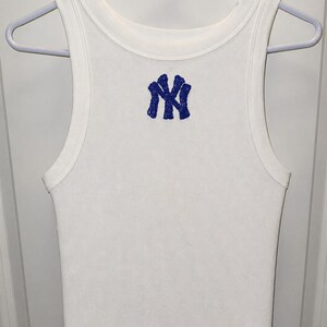 Könnte beinhalten: Weißes geripptes Tanktop mit blauem, gesticktem NY-Logo. Das ärmellose Oberteil hängt an einem weißen Kleiderbügel. Das Oberteil ist aus geripptem Stoff gefertigt.