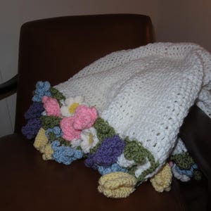 May include: A white crocheted blanket with a floral border in shades of pink, blue, yellow, and purple. The blanket is draped over a brown chair, showcasing the handmade details and soft texture. A cozy home decor item.