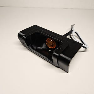May include: A black rectangular automotive light housing with a curved edge. An amber-coloured light bulb is visible inside the housing. White and black wires extend from the housing.