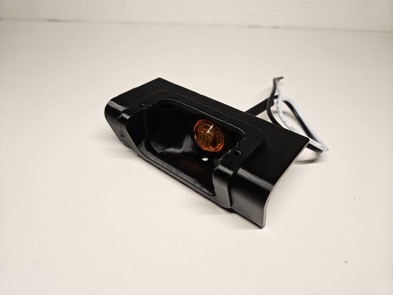 May include: A black rectangular automotive light housing with a curved edge. An amber-coloured light bulb is visible inside the housing. White and black wires extend from the housing.