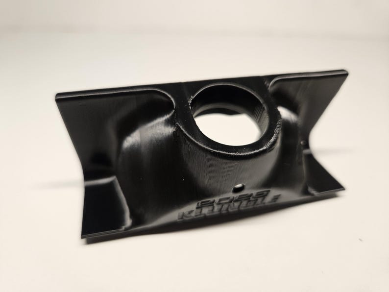 May include: A black, 3D-printed component with a central circular aperture and flared sides. The object has a textured finish, and the text "BOBS" is visible. The design is symmetrical, suggesting it's a functional part or a component.