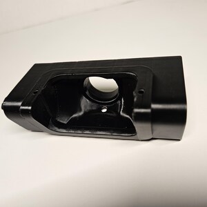 May include: A black plastic component with a rectangular body and a circular aperture. The object has a smooth, matte surface and a recessed interior. It appears to be a housing or bracket. The design includes rounded corners and an angled side.