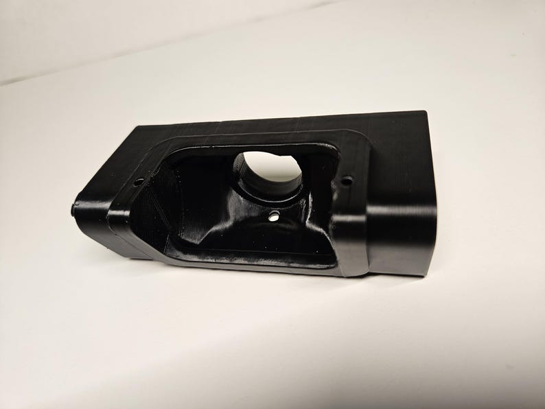 May include: A black plastic component with a rectangular body and a circular aperture. The object has a smooth, matte surface and a recessed interior. It appears to be a housing or bracket. The design includes rounded corners and an angled side.