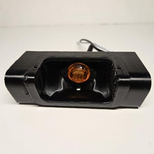 May include: A black rectangular automotive light unit with an amber bulb at its centre. The unit has a glossy finish and is set against a white background. Likely for a vehicle.