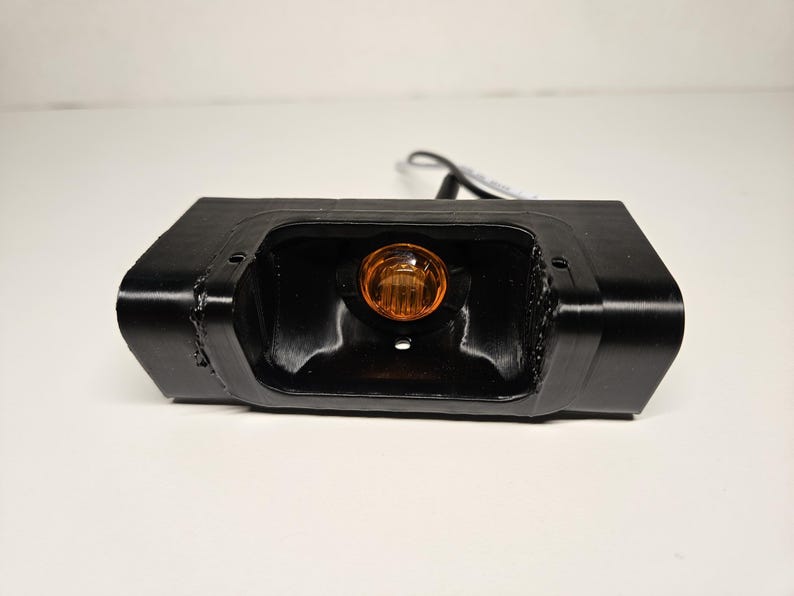 May include: A black rectangular automotive light unit with an amber bulb at its centre. The unit has a glossy finish and is set against a white background. Likely for a vehicle.