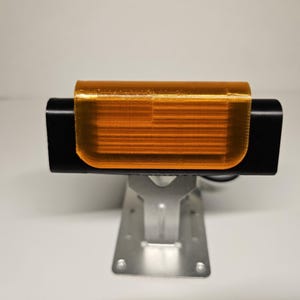 May include: A black rectangular device with an orange, translucent cover, mounted on a silver metal bracket. The device appears to be electronic, possibly a light or sensor, and is set against a plain white background.
