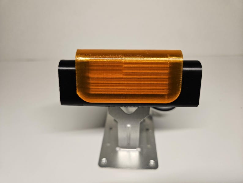 May include: A black rectangular device with an orange, translucent cover, mounted on a silver metal bracket. The device appears to be electronic, possibly a light or sensor, and is set against a plain white background.