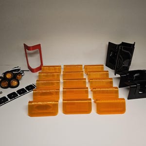 May include: A selection of automotive lighting components, featuring amber lenses, black housings, a red bracket, and a set of small round lights. Also included are mounting screws and a black mounting strip. The parts are arranged on a white surface.