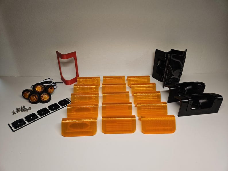 May include: A selection of automotive lighting components, featuring amber lenses, black housings, a red bracket, and a set of small round lights. Also included are mounting screws and a black mounting strip. The parts are arranged on a white surface.