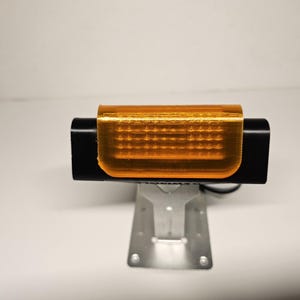 May include: A black rectangular device with an orange translucent cover, mounted on a silver metal stand. The device has a series of small, evenly spaced holes visible through the orange cover. The background is plain white.