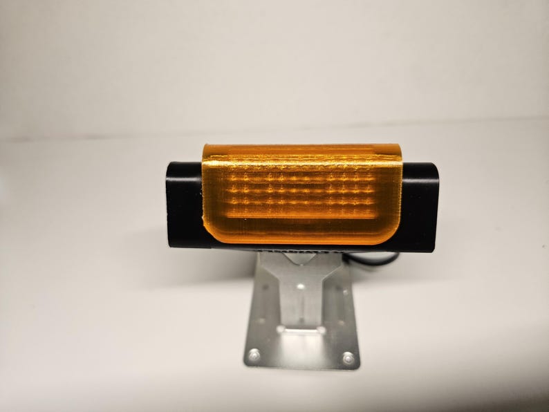 May include: A black rectangular device with an orange translucent cover, mounted on a silver metal stand. The device has a series of small, evenly spaced holes visible through the orange cover. The background is plain white.