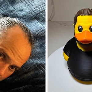 Puede incluir: Un pato de goma amarillo con un peinado casta&ntilde;o moldeado y barba negra, que viste una camiseta negra lisa. El pato tiene un pico naranja brillante y ojos negros, dise&ntilde;ado para parecerse a una persona con vello facial y ropa oscura.
