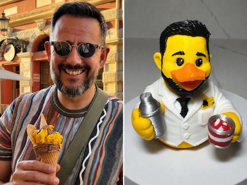 Puede incluir: Una figura de pato de goma amarillo caracterizado como camarero con pelo negro y barba, vistiendo una chaqueta blanca con corbata negra. El pato sostiene una coctelera plateada en un ala y un peque&ntilde;o cubo a rayas rojas y blancas en la otra.