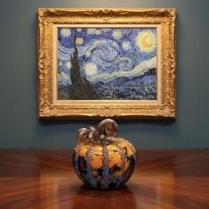 May include: A glass pumpkin sculpture featuring a rounded body with vertical ribs in shades of orange, blue, and black, topped with a clear, spiraled glass stem. The piece is displayed in front of a framed print of The Starry Night painting.