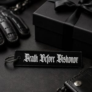 May include: A rectangular black fabric keychain featuring the white embroidered text Death Before Dishonor in a gothic font. The keychain includes a silver metal ring attached to one end, showcasing a clean, embroidered finish on the fabric surface.