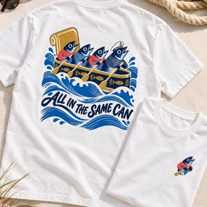 Może przedstawiać: Biały t-shirt z grafiką na plecach przedstawiającą cztery ryby wiosłujące w łodzi z napisem "ALL IN THE SAME CAN" w kolorze niebieskim oraz mniejszą, pasującą grafiką ryby na piersi. Wzór wykorzystuje kolory niebieski, czerwony i złoty.