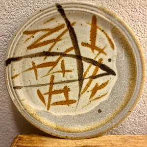 May include: A round ceramic plate with a speckled off-white base, featuring abstract brown and dark charcoal brushstroke patterns. The plate has a raised rim and is displayed with a US dime coin for scale, showing the relative size of the object.