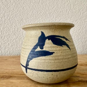 May include: A round, speckled beige ceramic pot with a wide, flared rim. The exterior features a hand-painted dark blue botanical design of leaves and a single horizontal line encircling the lower portion of the vessel's body.