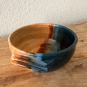 Op de afbeelding: Een ronde keramische kom met een tweekleurig glazuurontwerp. De ene kant van de kom is een warme, aardse beige kleur, terwijl de andere kant een diepe, glanzende blauwgroene kleur heeft. De twee kleuren vloeien in elkaar over en heeft oren.