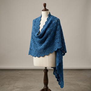 Könnte beinhalten: Ein blauer gehäkelter Schal, der auf einer Schaufensterpuppe präsentiert wird. Der Schal hat einen gewellten Rand und ein strukturiertes Muster. Die Farbe ist ein tiefes, sattes Blau, und der Schal ist drapiert, um sein Design zu zeigen.