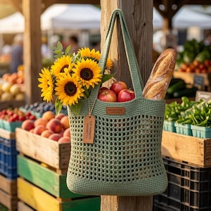 Könnte beinhalten: Eine hellgrüne gehäkelte Markt-Tragetasche gefüllt mit Sonnenblumen, roten Äpfeln und einem Baguette. Die Tasche hängt an einem Holzpfosten auf einem Bauernmarkt, mit Kisten voller Produkte im Hintergrund. Ein Etikett lautet "THE FARMERS MARKET CO."
