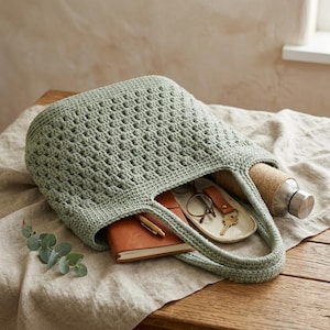Op de afbeelding: Een saliegroene, open geweven gehaakte tote bag gevuld met een bruin leren notitieboek, een pen, sleutels, een bril en een roestvrijstalen waterfles. De tas ligt op een neutraal gekleurde linnen doek.