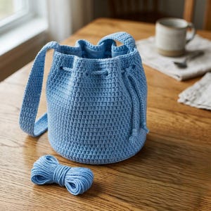 May include: A light blue crochet bucket bag with a single shoulder strap and a drawstring closure. The bag features a textured, woven stitch pattern throughout its cylindrical body. A small, coiled length of matching light blue yarn sits nearby.