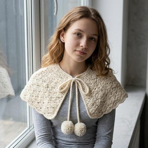 Puede incluir: Cuello de ganchillo color crema con borde festoneado y cierre de lazo. El cuello presenta dos pompones color crema colgando de la corbata. El cuello está hecho de un hilo texturizado y se usa sobre una camisa gris de manga larga.