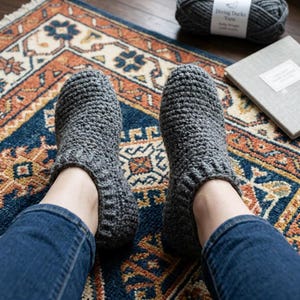 May include: A pair of gray crocheted slippers. The slippers are made of a thick yarn and have a ribbed cuff. The person is wearing blue jeans. The slippers are on a colorful rug with a geometric pattern. A ball of yarn and a book are in the background.