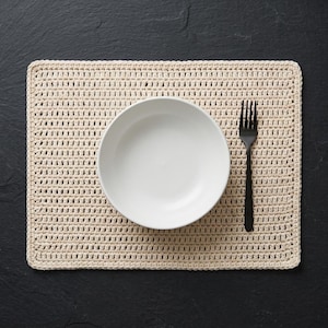 May include: A rectangular cream-colored crocheted placemat featuring a textured stitch pattern. Centered on the placemat is a plain white round bowl, and to the right of the bowl lies a single black metal fork with four tines.