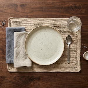 May include: A rectangular beige crocheted placemat featuring a speckled cream ceramic plate, a grey linen napkin, a beige linen napkin, a silver spoon, and a clear glass tumbler. The items are arranged neatly on a dark brown wooden surface.