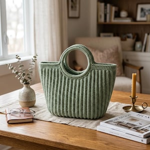 May include: A sage green, ribbed-knit tote bag with a circular handle. The bag sits on a wooden table with a neutral-toned runner. A small vase with greenery, a notebook, and a candle are also on the table.