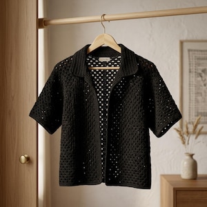 Puede incluir: Camisa negra de crochet de manga corta con cuello, colgada de una percha de madera. La camisa tiene un diseño de tejido abierto. La palabra "LOOM" es visible en el interior del cuello. La camisa se muestra sobre un fondo neutro.
