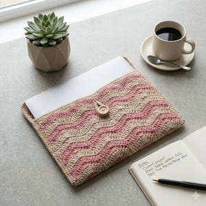May include: A handmade laptop sleeve with a pink and beige wave pattern. The crochet sleeve has a wooden button closure. A succulent in a geometric pot, a cup of coffee, and a notebook with notes are also on the table.
