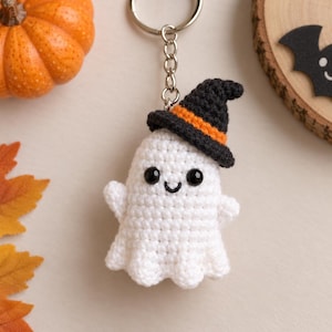 May include: A white crocheted ghost keychain featuring a black witch hat with an orange band. The ghost has two small black bead eyes and a stitched black smile. It is attached to a silver-colored metal keyring with a short chain link connector.