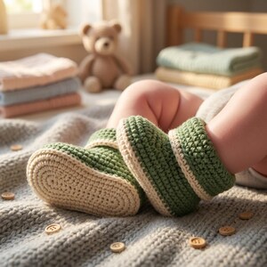 May include: Hand-crocheted baby booties in green and cream. The booties are on a gray blanket with wooden buttons. A teddy bear and folded blankets are in the background, suggesting a nursery setting. The booties are designed to keep baby's feet warm.