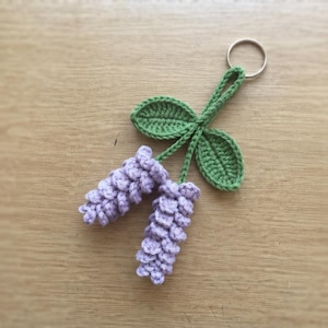 May include: A crochet keyring featuring two textured lavender-coloured flower clusters attached to a green stem with two green leaves. The floral design is connected to a silver-toned metal split ring at the top for attachment to keys or bags.
