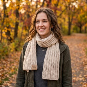May include: A beige, ribbed-knit scarf with a rectangular tag is draped around the neck. The scarf is worn over a gray sweater and a green jacket. The background features a blurred autumn scene with colorful foliage.