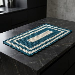 May include: A rectangular crochet mat featuring alternating concentric bands of deep teal and off-white yarn. The piece is constructed with a classic granny square stitch pattern and finished with a decorative scalloped edge border.
