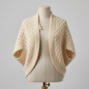 May include: Cream-colored, open-front crochet shrug with short sleeves. The handmade garment features a textured stitch pattern and a small tassel detail at the neckline. The shrug is displayed on a mannequin.