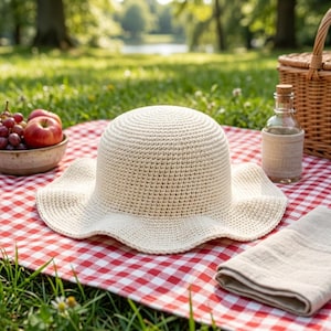 May include: A cream-coloured, crocheted sun hat with a wide, wavy brim rests on a red and white checked picnic blanket. The hat is next to a picnic basket, a bottle, a napkin, and a bowl of fruit, suggesting a relaxing outdoor scene.